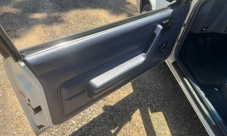 Driver's door is open and showing a blue door panel, black door handle, and black window crank.